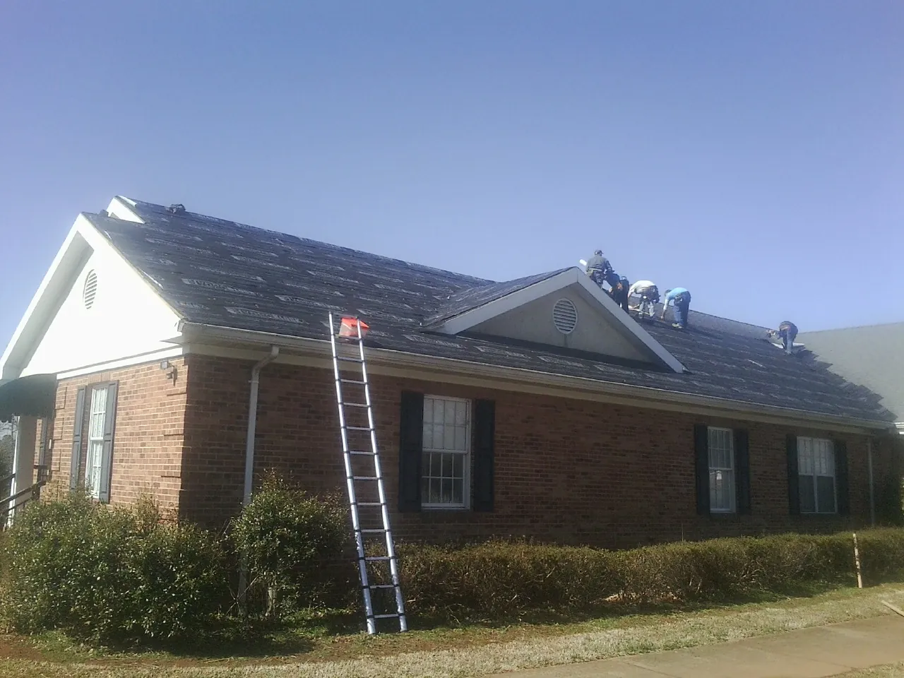 Crew tearing shingles off a brick ranch roof with a ladder set — Metal Roof Repair work by our crew in Luther