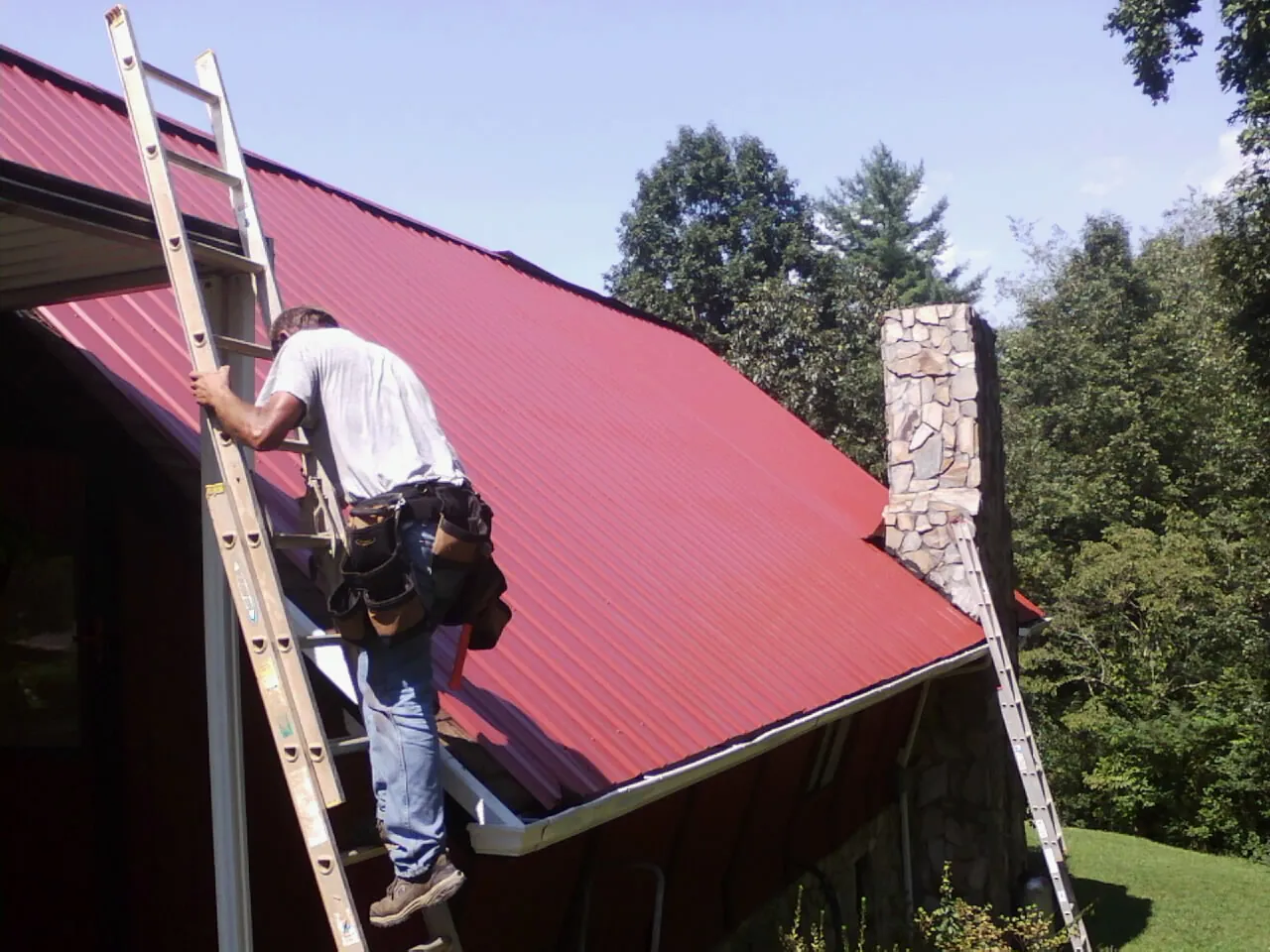 Roofer on a ladder beside a red metal roof and stone chimney — Soffit & Fascia Repair by Roofing Brothers in Luther